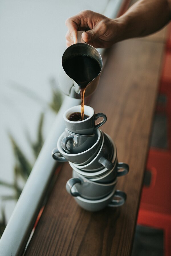 A hand pouring coffee into a tower of coffee and espresso cups. Photo by Nathan Dumlao on Unsplash.