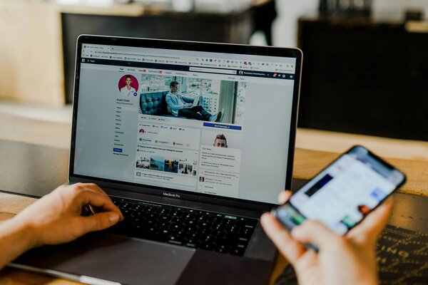 An open laptop showing a social media profile and a person with one hand on the keyboard and one on a mobile phone, with the screen on. Photo by Austin Distel on Unsplash.