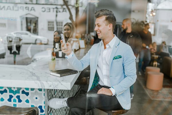 Photo of a young man sitting at a bar, holding up and looking at his phone and smiling. Reflection of two yougn women smiling in the window. Photo by Austin Distel on Unsplash.