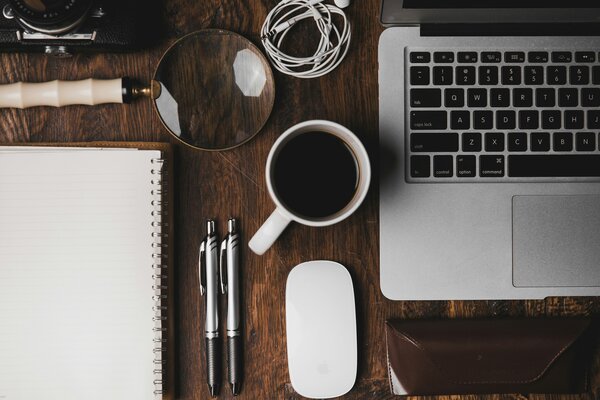 Top down view of a wooden desk, notepad, pens, magnifying glass, coffee, keyboard, mouse, cable, camera, etc. Photo by ian dooley on Unsplash