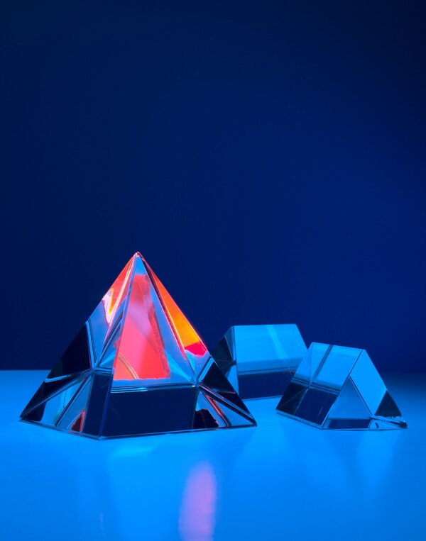 A glass pyramid that is bigger and reflecting pink, orange and yellow colours. A light blue surface with two smaller pyramids on it (only in shades of blue). A dark blue backdrop and the three pyramids are placed on a light blue surface. Photo by Michael Dziedzic on Unsplash.